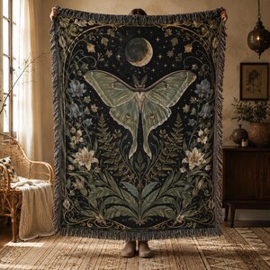 May include: A rectangular woven throw blanket featuring a large central luna moth in shades of pale green against a dark black background. The design includes a crescent moon, stars, and detailed botanical illustrations of lilies and ferns.