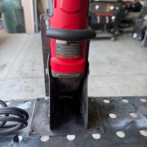 Puede incluir: Una amoladora angular roja y negra montada verticalmente en un soporte de acero hecho a medida. La herramienta cuenta con un mango lateral negro y una etiqueta de advertencia con texto. El soporte tiene una placa base plana con cuatro orificios.