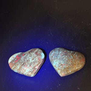 May include: Two heart-shaped stones featuring a mottled texture with a blend of earthy brown, vibrant turquoise, and bright red mineral inclusions. The stones are displayed against a dark background under a blue light source.
