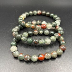 May include: Two elastic bracelets composed of round, polished beads in shades of forest green, moss green, translucent white, and opaque red. The beads feature natural mottled patterns and variations in color, creating a textured appearance.