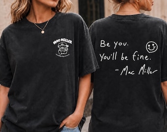 May include: A charcoal black short-sleeve t-shirt featuring a small white graphic on the front chest with the text Mac Miller. The back displays the white text Be you. You'll be fine. - Mac Miller and a simple smiley face icon.
