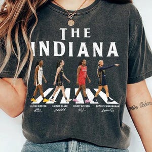 May include: A charcoal gray t-shirt featuring the text THE INDIANA in white capital letters. Below, four basketball players are depicted in action, with their names Aliyah Boston, Caitlin Clark, Kelsey Mitchell, and Sophie Cunningham printed below.
