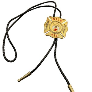 May include: A bolo tie featuring a gold-toned metal emblem shaped like a Maltese cross. The emblem displays the text NILE and 111 in red, with a central white circle containing a red star. The tie has a black braided cord with gold metal tips.