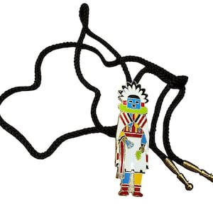 May include: A bolo tie featuring a central metal pendant shaped like a stylized figure wearing a white headdress, a white robe with red and blue stripes, and yellow leggings. The pendant is attached to a black braided cord with gold metal tips.