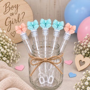 May include: Five clear plastic bubble wands with bow-shaped toppers in alternating pastel peach and light blue colors, standing upright in a glass mason jar wrapped with a brown twine bow. The wands feature a long stem and a rectangular loop.