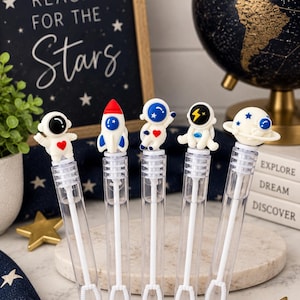 May include: Five clear plastic bubble wands featuring space-themed toppers. The toppers include an astronaut with a red heart, a white rocket ship, an astronaut waving, an astronaut with a yellow lightning bolt, and a planet with a ring.