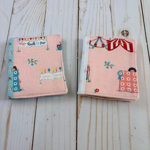 May include: Two rectangular fabric-covered notebooks featuring a light pink background with a carnival theme. The print includes a ferris wheel, a red and white striped tent, and signs reading Welcome to the Quilt Fair and Cotton Candy.