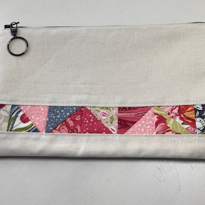 May include: A rectangular, cream-colored zippered pouch with a decorative band of colorful, triangular fabric patches. The patches feature floral and geometric patterns in shades of pink, red, blue, and green. A black zipper pull with a ring is visible.