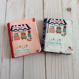 May include: Two rectangular fabric-covered notebooks featuring a printed design of a shop stall with the text Vintage Quilts and Cotton Candy. The left notebook has a pink background with a red spine, while the right has a white background with a blue checkered spine.