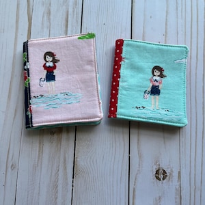 May include: Two fabric-covered small notebooks. One is pink with a floral and navy blue border, the other is turquoise with a red polka dot border. Each features an embroidered image of a girl standing in water, holding a bag.