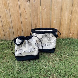 May include: Two white drawstring bags with black trim and a black and white pattern of circles. The bags are sitting on a green grassy surface.
