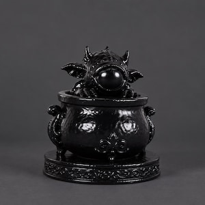 May include: A glossy black decorative container featuring a sculpted creature with horns and large eyes emerging from the top of a rounded cauldron-shaped vessel. The base is circular with intricate embossed detailing around the edge.