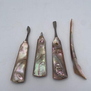 May include: A set of four manicure tools featuring iridescent abalone shell handles with natural pearlescent patterns in shades of pink, green, and cream. The set includes three metal-tipped implements and one curved shell cuticle pusher.