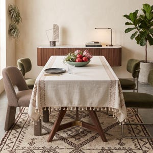 May include: A rectangular off-white tablecloth featuring a tan geometric border pattern and small hanging tassels along the edges. The fabric has a visible woven texture, draped over a wooden dining table with a dark brown finish and metal legs.