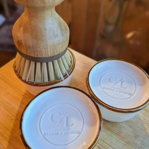 May include: A light-colored wooden dish brush with stiff bristles sits inside a white ceramic holder. Two additional white ceramic holders with brown rims are present, each featuring a circular embossed logo with the letters C/L and text.