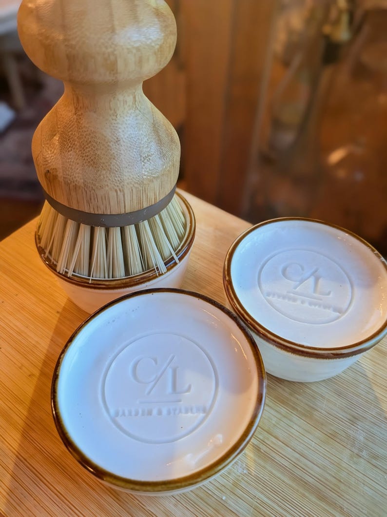 May include: A light-colored wooden dish brush with stiff bristles sits inside a white ceramic holder. Two additional white ceramic holders with brown rims are present, each featuring a circular embossed logo with the letters C/L and text.