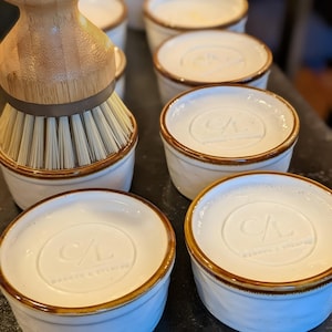 May include: A bamboo dish brush with stiff bristles sits next to several white ceramic ramekins. Each ramekin features a brown rim and a circular embossed logo on the top surface that reads CL and includes the text WARETO & EVELYN.