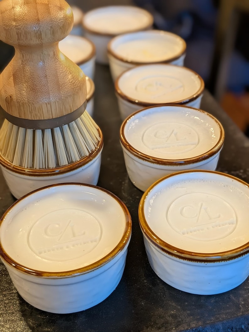 May include: A bamboo dish brush with stiff bristles sits next to several white ceramic ramekins. Each ramekin features a brown rim and a circular embossed logo on the top surface that reads CL and includes the text WARETO & EVELYN.