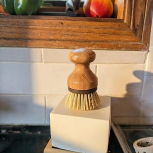 May include: A round wooden dish brush with a light brown handle and stiff, light-colored bristles. The brush features a dark brown ring connecting the handle to the bristles and is positioned upright on a solid white block resting on a wooden base.