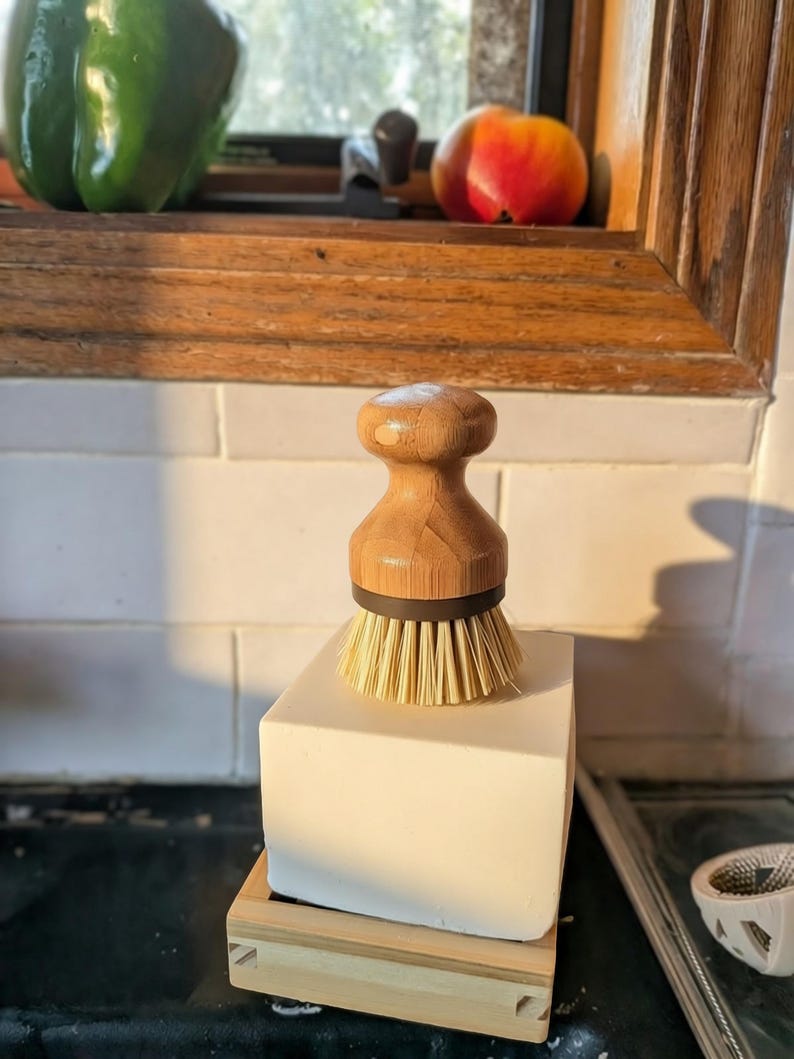 May include: A round wooden dish brush with a light brown handle and stiff, light-colored bristles. The brush features a dark brown ring connecting the handle to the bristles and is positioned upright on a solid white block resting on a wooden base.