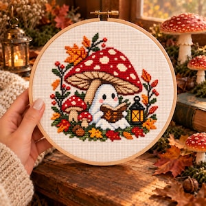 Può includere: Un telaio da ricamo circolare in legno contenente un motivo a punto croce di un fantasma bianco che legge un libro sotto un grande fungo rosso a pois bianchi, circondato da foglie autunnali, funghi più piccoli e una lanterna.