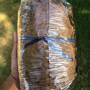 Puede incluir: Un paquete rectangular de hojas marrones secas envuelto en una película de plástico transparente. El paquete está asegurado en el centro con una cinta delgada de color azul oscuro atada en un lazo simple. Las hojas tienen una textura.