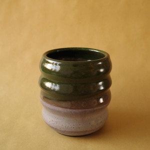 May include: A ceramic cup featuring a two-tone glaze with a glossy dark forest green upper section and a speckled light lavender lower section. The vessel has a cylindrical shape with three distinct horizontal ridges along the body.
