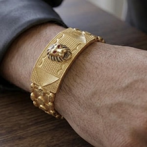 May include: A wide gold-toned metal cuff bracelet featuring a central lion head emblem with red eyes. The surface of the bracelet has a textured, patterned finish with geometric details and a linked chain-style design along the edges.