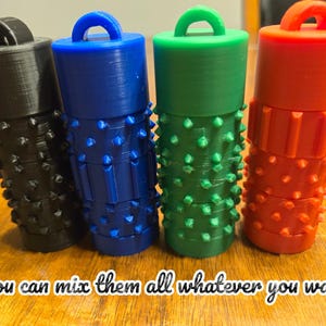Peut inclure: Quatre récipients cylindriques en noir, bleu, vert et orange, chacun doté d'une boucle supérieure et d'une surface texturée avec de petites pointes et des crêtes verticales. Le texte "You can mix them all whatever you want" est imprimé en bas.
