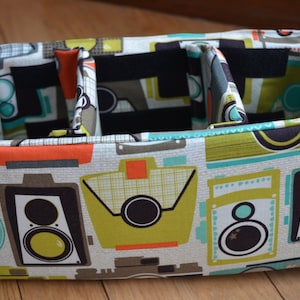 May include: A fabric storage box with three compartments. The box is decorated with a retro pattern of cameras and music speakers in shades of green, blue, orange, and brown.