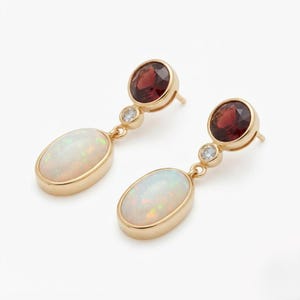 May include: A pair of gold drop earrings featuring a round, deep red garnet stone at the top, a small clear accent stone in the middle, and a large, oval-shaped iridescent opal at the bottom, all set in polished yellow gold metal settings.