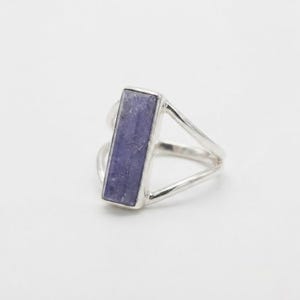 May include: A silver ring featuring a rectangular, vertically oriented, light purple gemstone set in a smooth metal bezel. The band is constructed from two thin, polished silver wires that diverge from the base to meet the gemstone setting.
