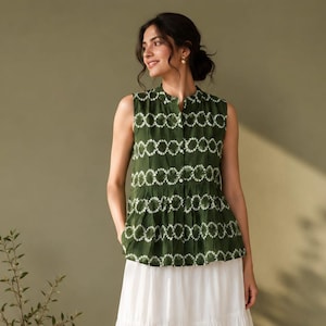 Puede incluir: Una blusa sin mangas de color verde oliva con un patrón circular de teñido anudado en blanco, cierre frontal de botones y cintura plisada, combinada con una falda larga blanca de niveles. El conjunto se completa con sandalias claras.