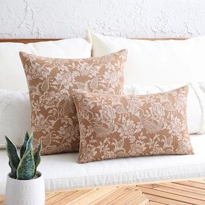 May include: Two throw pillows featuring a tan background with a white floral and paisley pattern. One pillow is square and the other is rectangular. Both pillows are displayed against a white background with a small green plant in the foreground.