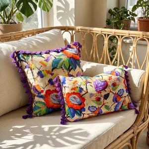 Puede incluir: Dos cojines decorativos con una base de color crema y bordados florales y de hojas multicolores vibrantes. Cada cojín está rematado con borlas moradas en los bordes, destacando hilos de colores naranja, azul, verde y amarillo brillante.