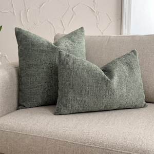 Puede incluir: Dos cojines decorativos de color verde salvia con un acabado de tela tejida texturizada. Un cojín es de forma cuadrada y está colocado en posición vertical, mientras que el otro es rectangular y está situado delante, ambos con un patrón de color verde moteado.