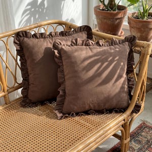 May include: Two square throw pillows in a solid dark brown color. Each pillow features a decorative ruffled edge trim made from the same fabric. The pillows are positioned on a light-colored woven cane bench with a curved wooden frame structure.
