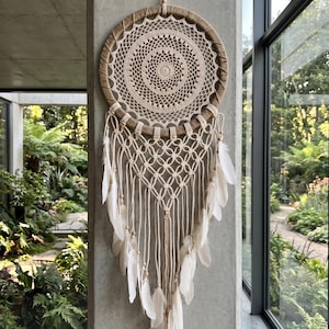 May include: A circular dream catcher featuring a tan jute-wrapped hoop with a central cream-coloured crocheted mandala pattern. Below the hoop, cream-coloured macrame cord is knotted in a diamond pattern, accented with small wooden beads and white feathers.
