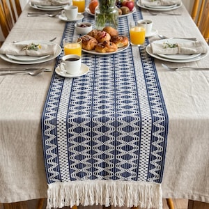 Puede incluir: Un camino de mesa rectangular de tela azul intenso con un patrón geométrico de diamantes blancos y rayas verticales, acabado con largos flecos de color crema en ambos extremos, extendido sobre un mantel de lino de tono neutro.