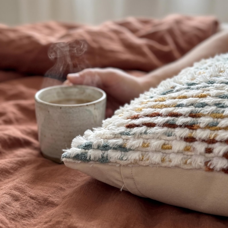 Puede incluir: Un coj&iacute;n decorativo texturizado con un patr&oacute;n tejido de hilos blancos, amarillo mostaza, verde azulado y color &oacute;xido. El coj&iacute;n tiene una base de tela beige lisa y est&aacute; colocado junto a una taza de cer&aacute;mica con una bebida caliente humeante.