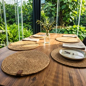 May include: A set of round, woven jute placemats arranged on a wooden dining table. Each placemat features a spiral pattern and a small brown leather-like tag. A clear glass vase with green olive branches sits in the center with ceramic plates.