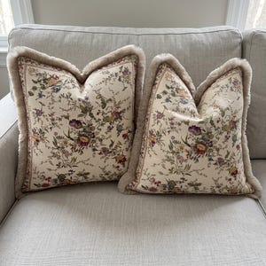 May include: Two square throw pillows with a cream-colored base featuring a multi-colored floral pattern of red, purple, yellow, and green blossoms. Each pillow is finished with a thick, soft, light brown faux fur trim along the outer edges.