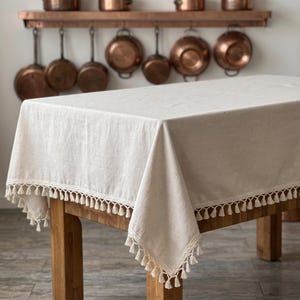 Pode incluir: Uma toalha de mesa retangular em tom branco sujo com acabamento em tecido texturizado, apresentando uma borda decorativa de crochê ao longo das laterais, adornada com pequenas borlas pendentes. A toalha cobre uma mesa de madeira com veios.