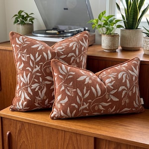 May include: Two rectangular throw pillows in a burnt orange fabric featuring a white and light brown botanical leaf and fruit pattern. The pillows have matching orange piping along the edges and a textured finish, displayed as a pair on a wooden surface.