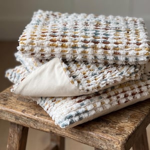 Puede incluir: Una pila de tres cojines cuadrados con tela blanca texturizada y copetuda, con un patr&oacute;n repetitivo de peque&ntilde;os puntos en relieve en tonos amarillo mostaza, naranja &oacute;xido y azul apagado. La parte inferior de los cojines es de color crema liso.