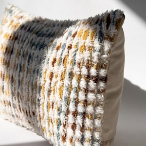 Puede incluir: Un coj&iacute;n rectangular con un panel frontal texturizado y peludo que presenta un patr&oacute;n de rayas verticales en tonos crema, amarillo mostaza, marr&oacute;n &oacute;xido y azul apagado. El panel lateral est&aacute; hecho de una tela lisa de color crema.