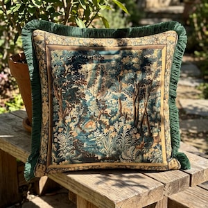 Puede incluir: Un cojín cuadrado con un diseño de tapiz tejido que muestra un paisaje forestal con árboles, follaje y un pequeño animal. El cojín está bordeado por un marco decorativo de color tostado y rematado con un fleco de color verde oscuro en los cuatro lados.