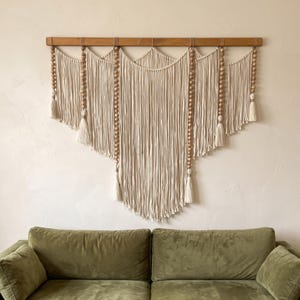 May include: A large macrame wall hanging suspended from a horizontal wooden rod. The piece features off-white cotton cord arranged in a layered, scalloped pattern with vertical strands of natural wooden beads ending in decorative tassels.
