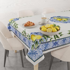 Könnte beinhalten: Eine rechteckige Tischdecke mit weißer Mitte und einem blau-weißen Blumenrand mit gelben Zitronen- und grünen Blattmotiven. Die Tischdecke liegt auf einem Tisch mit vier Stühlen, einer Schale mit Zitronen, einem Croissant und einer Kaffeetasse.