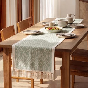 May include: A light green and white table runner with a repeating damask pattern and fringe trim on the ends, laid across a wooden dining table set with ceramic plates, bowls, a pitcher, and a bowl of salad with colorful vegetables.